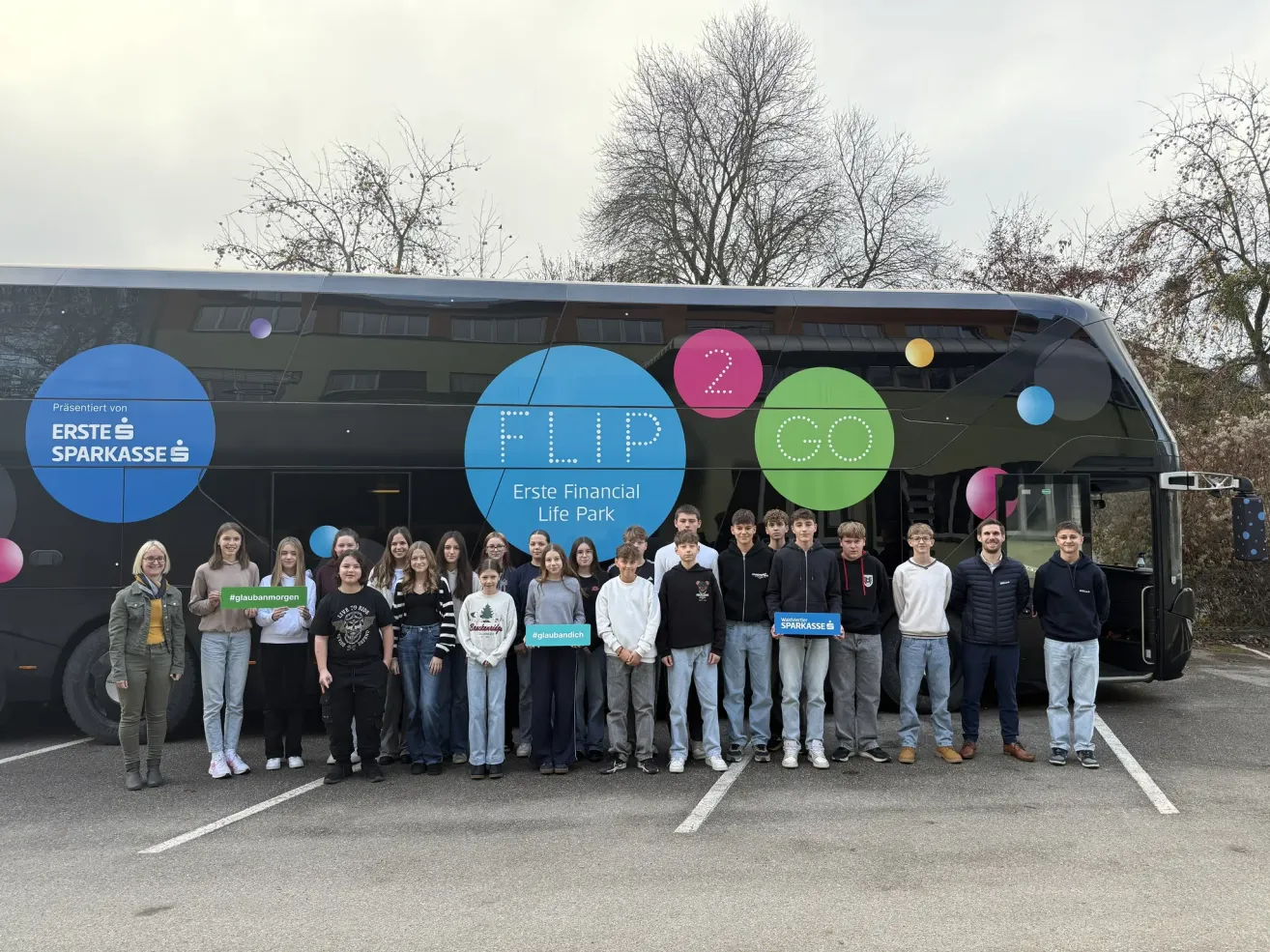 Eine Gruppe junger Menschen posiert vor einem Bus mit FLIP und 2 GO darauf geschrieben. Sie halten Schilder in den Haenden und tragen Freizeitkleidung.