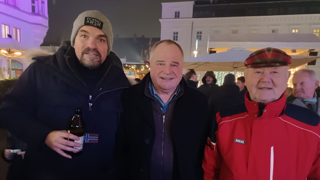 Drei Männer stehen draußen, zwei lächeln, einer hält eine Bierflasche. Sie tragen Winterkleidung, einer trägt einen roten Mantel. Hinter ihnen steht ein Gebäude mit Lichtern und einem Schirm.