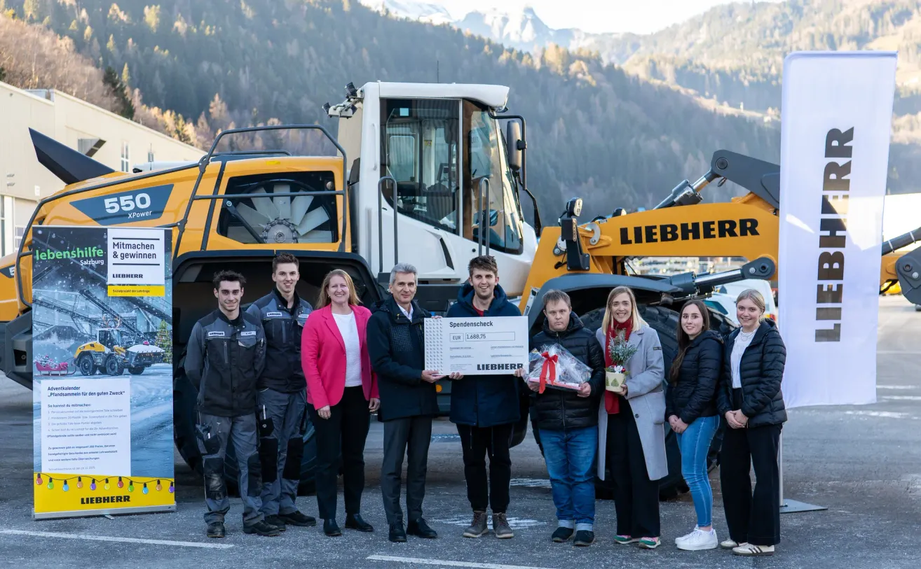Mehrere Personen stehen vor einem Liebherr-Baufahrzeug. Ein Mann hält einen Spendenscheck und ein Geschenk. Eine Frau hält eine Topfpflanze mit einem Band. Dahinter sind Berge.