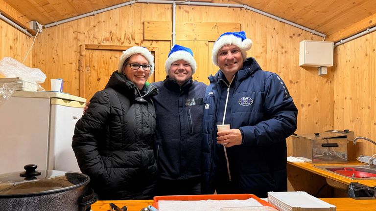 Drei Menschen in Winterkleidung, die in einem Raum mit Holzwänden lächelnd für ein Foto posieren, tragen Weihnachtsmützen und einer hält einen Becher.