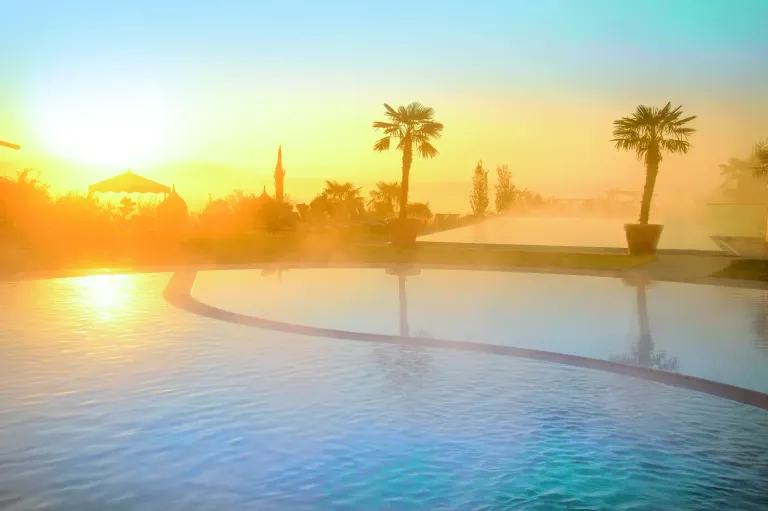Ein ruhiges Bild eines Schwimmbeckens mit einem Sonnenuntergang im Hintergrund. Palmen und Topfpflanzen umgeben das Becken und schaffen eine friedliche Atmosphäre.