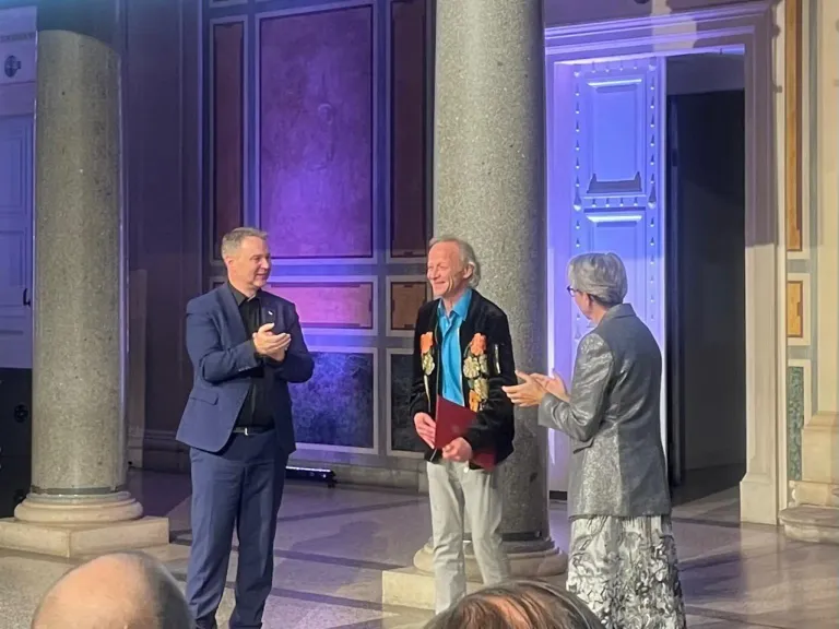 Drei Personen stehen auf einer Bühne, eine hält ein rotes Buch, während die anderen klatschen. Sie stehen vor einer Säule mit lila Beleuchtung.