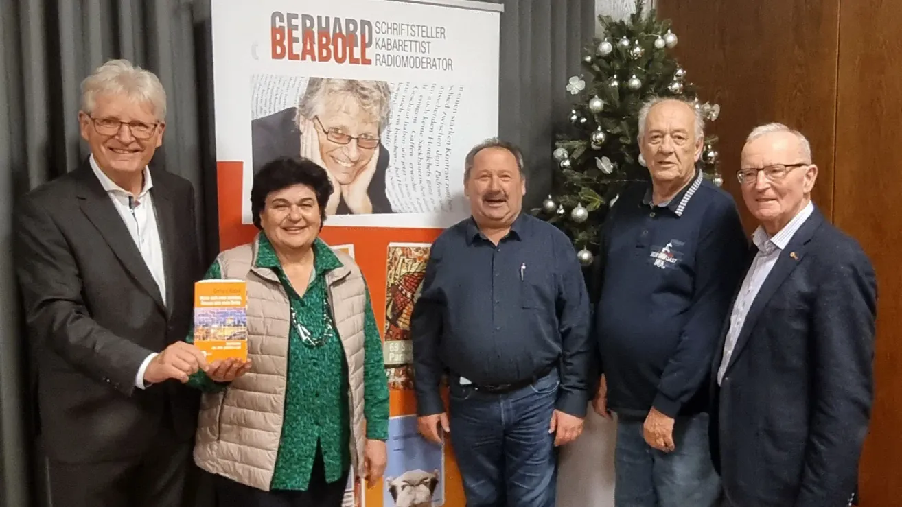 Drei Personen stehen vor einem Banner mit dem Text 'Gerhard Beabolle Schriftsteller Kabarettist Radiomoderator'. Eine Frau hält ein Buch, und ein Mann neben ihr lächelt. Dahinter steht ein geschmückter Weihnachtsbaum.