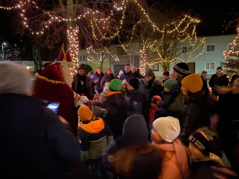 Bei Nacht versammelt sich eine Menschenmenge um einen Weihnachtsmann-Imitator. Die Szene ist mit Lichtern geschmückt und Gebäude sind im Hintergrund sichtbar.