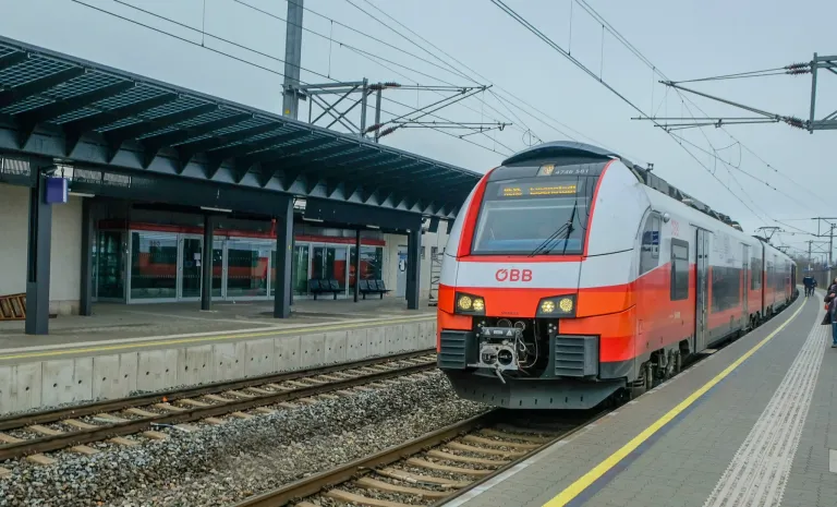Ein orange-weißer Zug mit den Buchstaben OBB auf der Vorderseite steht auf einem Bahngleis in der Nähe eines Bahnsteigs.