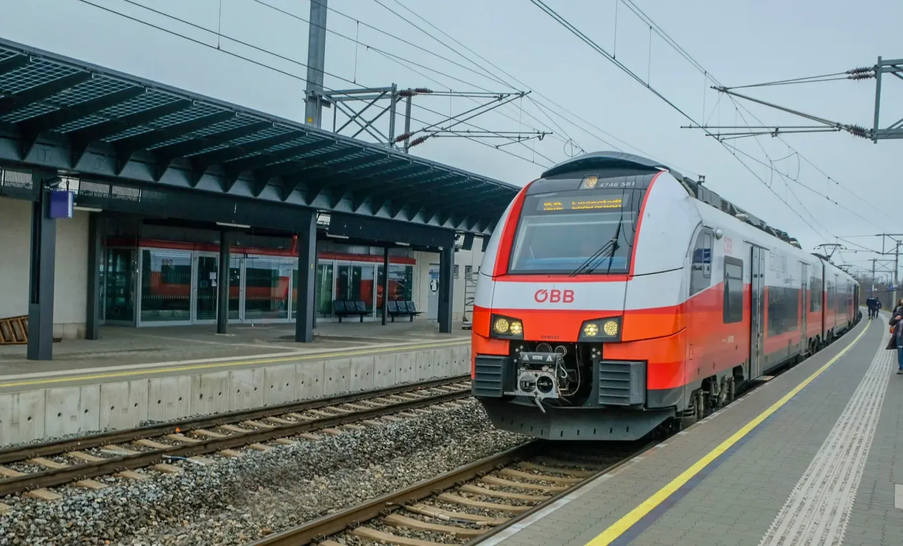 Ein orange-weißer Zug mit den Buchstaben OBB auf der Vorderseite steht auf einem Bahngleis in der Nähe eines Bahnsteigs.