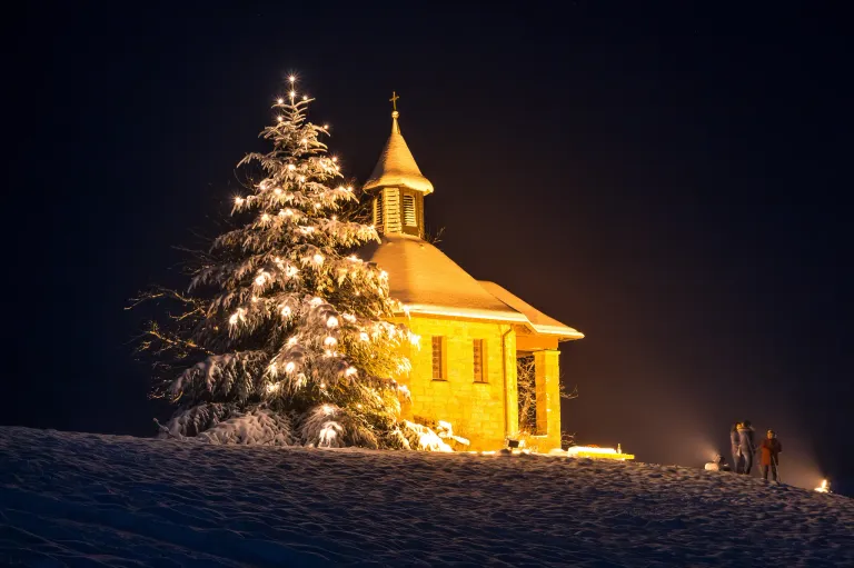 Ein beleuchteter Weihnachtsbaum steht vor einer verschneiten Kapelle. Die Kapelle hat einen Turm und Fenster, die beleuchtet sind.