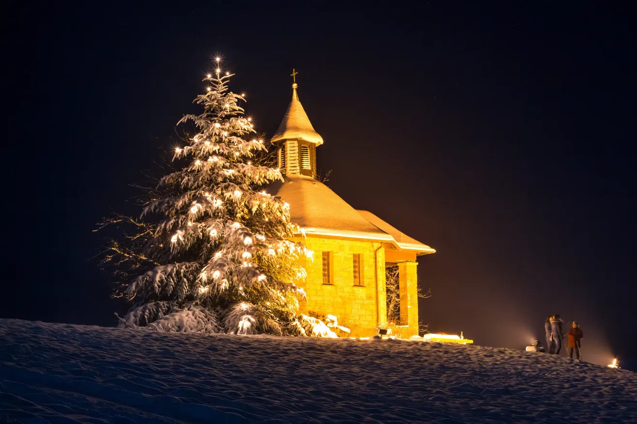 Ein beleuchteter Weihnachtsbaum steht vor einer verschneiten Kapelle. Die Kapelle hat einen Turm und Fenster, die beleuchtet sind.