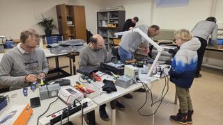 Eine Gruppe von Menschen arbeitet in einem Raum an elektronischen Geräten. Sie tragen Brillen und Stiefel. Es gibt Regale und einen Heizkörper im Raum.