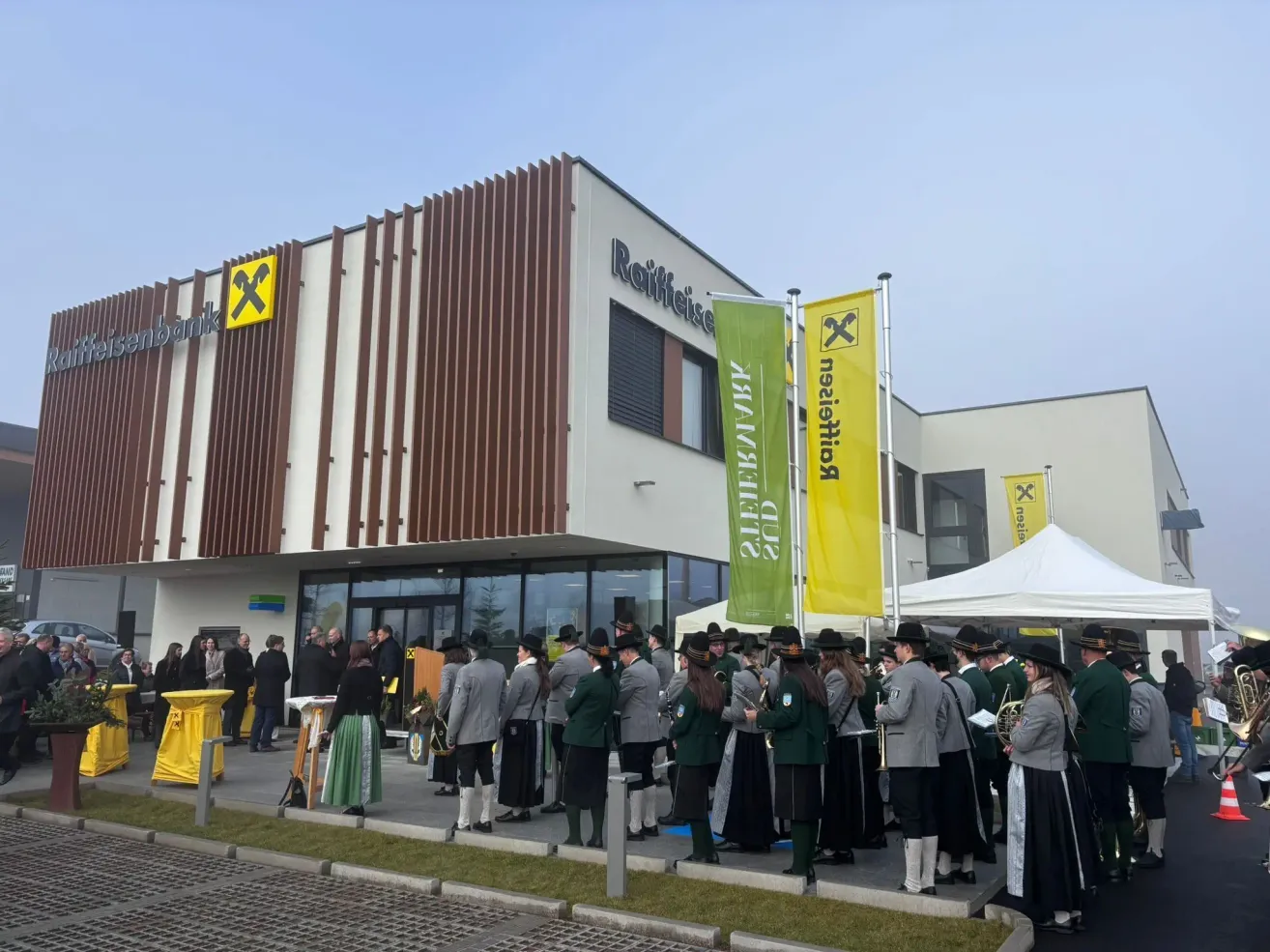 Eine Gruppe von Menschen in traditioneller Kleidung steht vor einem Gebäude mit einem Schild, auf dem Raiffeisenbank steht. Eine gelbe Fahne und ein weißes Zelt sind vorhanden.