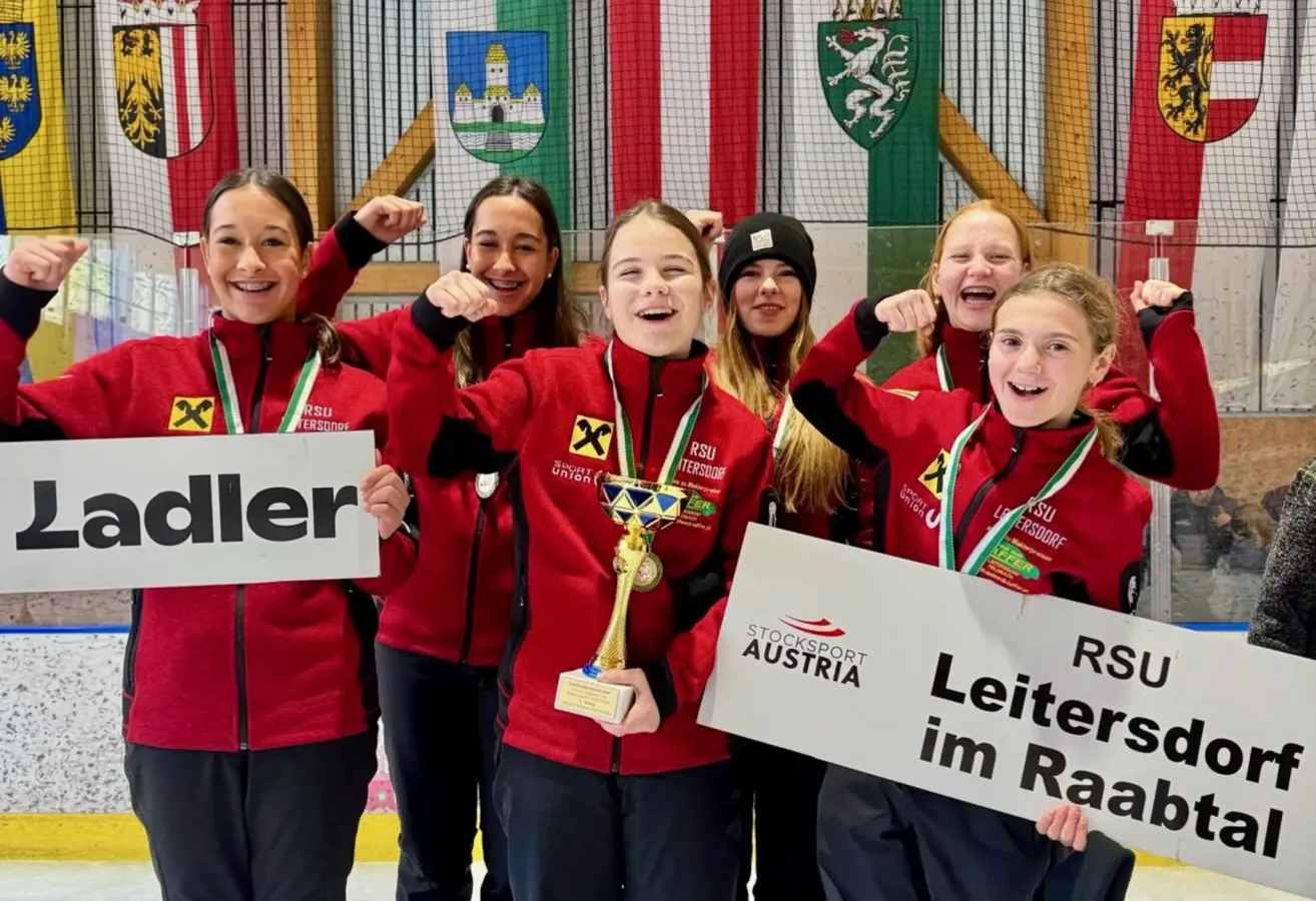 Eine Gruppe von Frauen in roten Trikots feiert einen Sieg. Sie halten Trophäen und Schilder mit den Worten 'Stocksport Austria'. Dahinter befinden sich Fahnen und ein Logo an einer Holzwand.