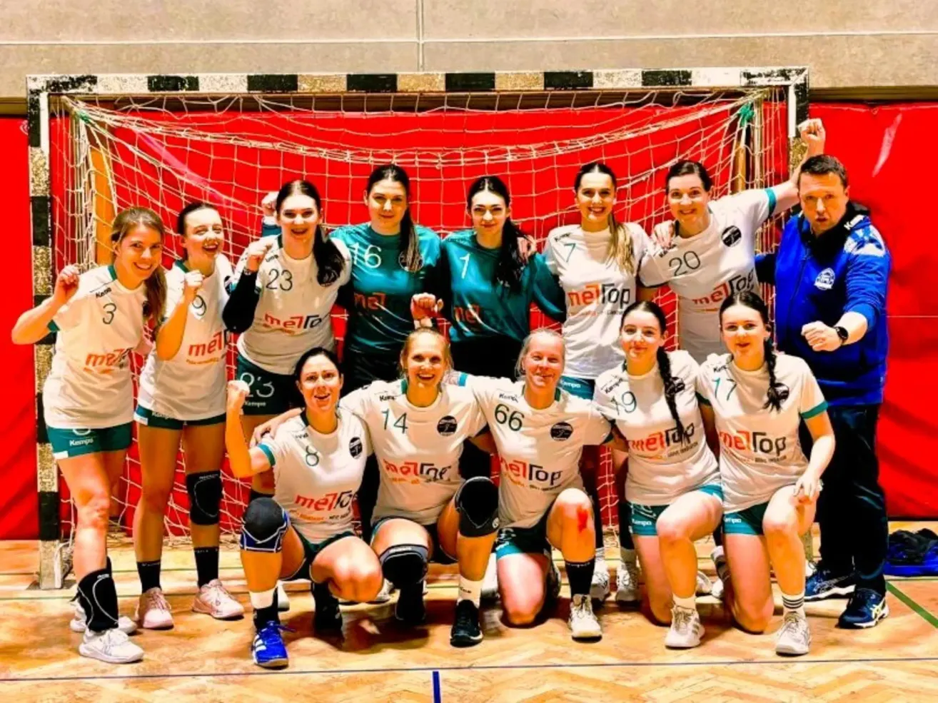 Eine Gruppe von Handballspielerinnen posiert für ein Foto mit ihren Teamtrikots, lächelt und hebt ihre Hände vor einem roten Tornetz.