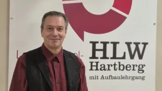 Ein lächelnder Mann steht vor einem Schild mit einem roten Kreis und dem Text HLW Hartberg. Er trägt ein weinrotes Hemd und eine schwarze Weste.