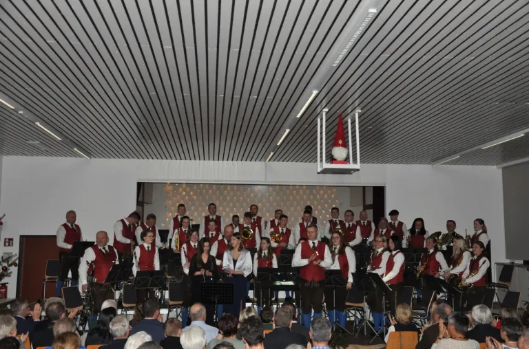 Ein großes Blasorchester spielt auf der Bühne in einer großen Veranstaltungshalle, mit Musikern in roten Westen und blauen Hosen. Ein Publikum sitzt vor ihnen. Eine Weihnachtsdekoration hängt von der Decke.