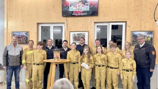 Eine Gruppe junger Leute in gelben Uniformen steht neben einem Tisch mit einem Laternenlicht. Sie befinden sich in einem Raum mit Holzwänden und Glastüren. Ein Foto ist an der Wand über ihnen montiert.