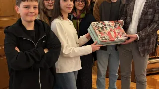 Eine Gruppe von Menschen steht in einem Raum. Eine Frau hält eine Geschenkbox. Der Junge auf der linken Seite hat die Arme verschränkt. Ein anderer Mann lächelt. Die anderen Leute schauen sie an.