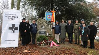 Eine Gruppe von Menschen steht an einem Denkmal, geschmückt mit Kränzen und einer Flagge, unter einem Baum mit einer Steintafel. Sie tragen Uniformen und Zivilkleidung.