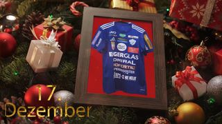 Ein gerahmtes Trikot mit Sponsorenlogos, das auf einem Weihnachtsbaum ausgestellt ist. Das Trikot ist blau mit weißen Texten. Unter dem Rahmen steht das Datum '17. Dezember'. Geschenke umgeben den Rahmen.