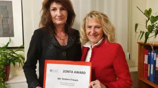 Zwei Frauen stehen in einem Innenraum zusammen. Die Frau auf der linken Seite hält ein Zertifikat. Das Zertifikat lautet 'Zonta Award' und 'Wir fördern Frauen'. Die Frau auf der rechten Seite lächelt und schaut in die Kamera.