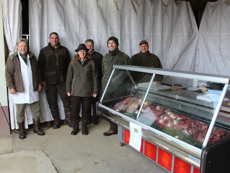 Eine Gruppe von Menschen in Jacken und Stiefeln steht vor einer gekühlten Fleischtheke. Sie lächeln und posieren für ein Foto.
