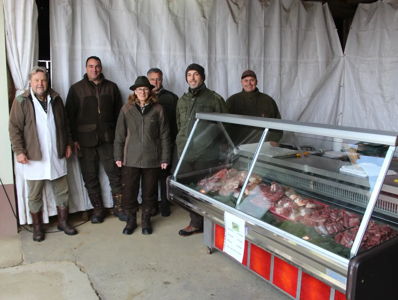 Eine Gruppe von Menschen in Jacken und Stiefeln steht vor einer gekühlten Fleischtheke. Sie lächeln und posieren für ein Foto.