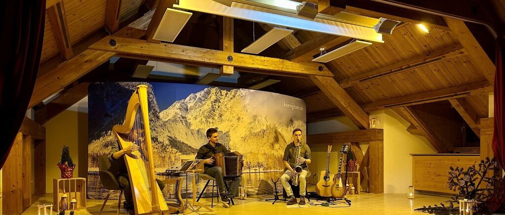 Drei Musiker spielen in einem Raum mit Holzbalken und Wänden. Die Person auf der linken Seite spielt eine Harfe, die mittlere spielt ein Akkordeon und die rechte eine Saxophon. Ein großes Bild eines Berges steht hinter ihnen.