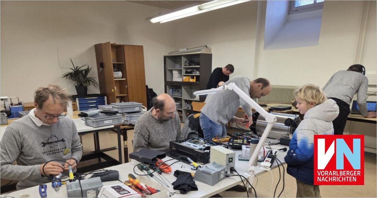 Mehrere Personen arbeiten in einer Werkstatt. Ein Mann steht und schaut sich ein Gerät an, während ein anderer Mann sitzt und ein Werkzeug in der Hand hält. Der Tisch hat verschiedene Werkzeuge, Kabel und elektronische Geräte. Hinter ihnen befinden sich Regale und Schränke mit Gegenständen.