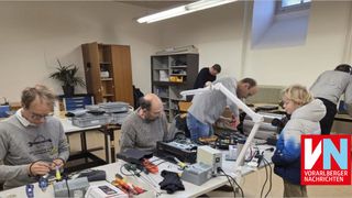 Mehrere Personen arbeiten in einer Werkstatt. Ein Mann steht und schaut sich ein Gerät an, während ein anderer Mann sitzt und ein Werkzeug in der Hand hält. Der Tisch hat verschiedene Werkzeuge, Kabel und elektronische Geräte. Hinter ihnen befinden sich Regale und Schränke mit Gegenständen.