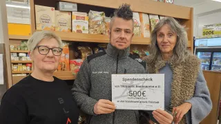 Drei Personen stehen vor einem Regal mit verschiedenen Lebensmitteln. Der Mann in der Mitte hält einen Spendenscheck über 500 Euro.