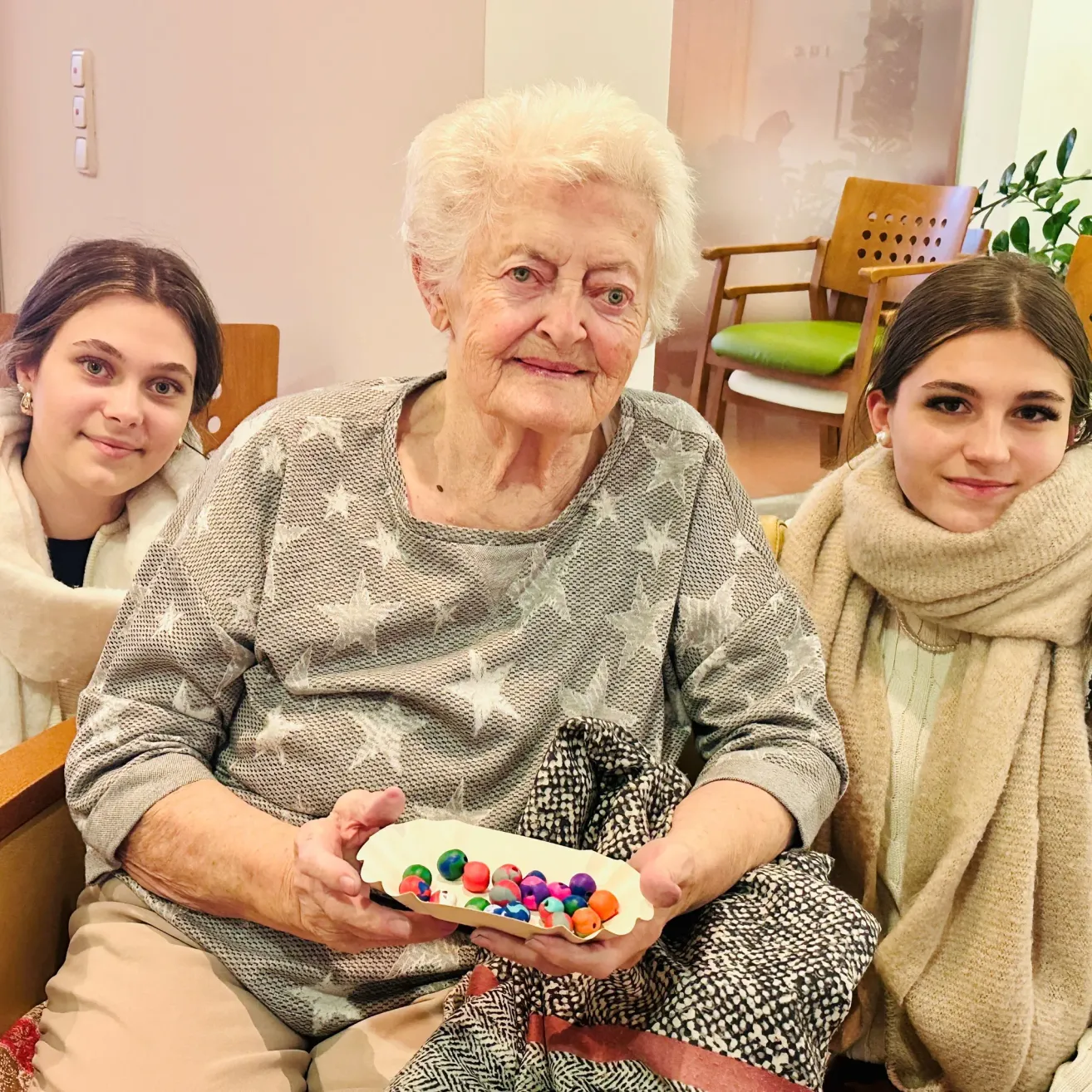 Eine ältere Frau, die eine bunte Perlenschale hält, lächelt mit zwei jungen Frauen, eine trägt eine weiße Jacke und die andere einen Schal. Sie sind in einem Raum mit einer weißen Wand und einer grünen Pflanze.
