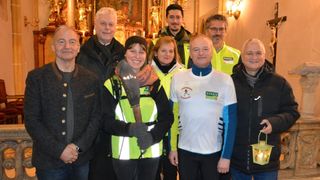 Eine Gruppe von Menschen in reflektierenden Westen steht in einer Kirche und posiert für ein Foto. Eine Frau hält eine Fackel, und ein Mann neben ihr trägt ein weißes Shirt mit 'KAUSCH' darauf.