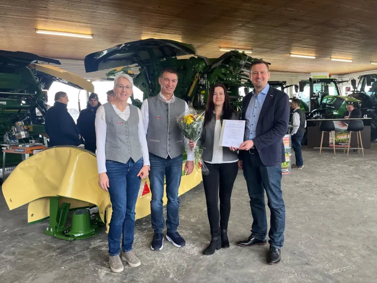 Drei Personen stehen vor einem grünen Traktor. Die Frau in der Mitte hält Blumen. Der Mann auf der rechten Seite hält ein Zertifikat. Die anderen beiden Personen lächeln in die Kamera.