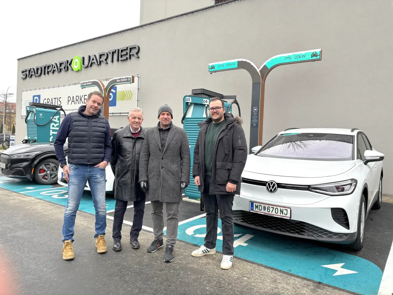 Vier Männer stehen neben einem weißen Volkswagen-Auto mit dem Kennzeichen MD 670 NJ an einer Ladestation mit dem Park Quartier-Schild an der Wand.