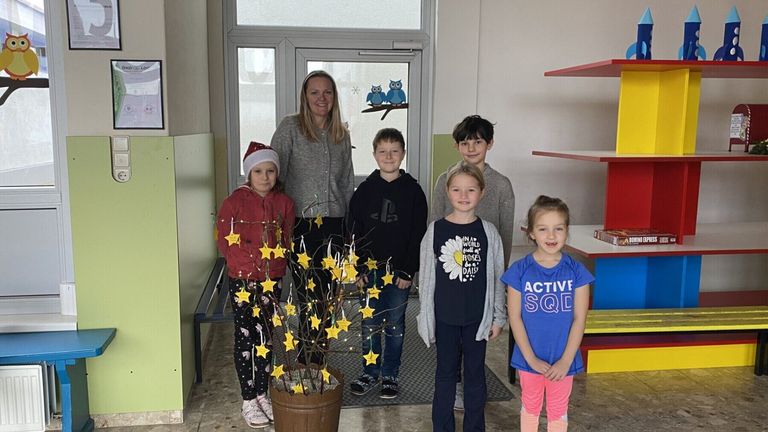 Eine Gruppe von Kindern und einer Frau steht in der Nähe einer Tür mit einem Zweig mit gelben Sternen und einem Topfpflanzen.