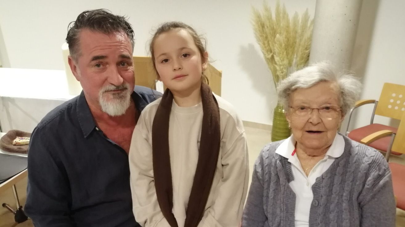 Drei Personen posieren für ein Bild in einem Raum. Ein Mädchen steht in der Mitte, und ein älterer Mann und eine ältere Frau sind auf beiden Seiten von ihr. Sie alle lächeln.