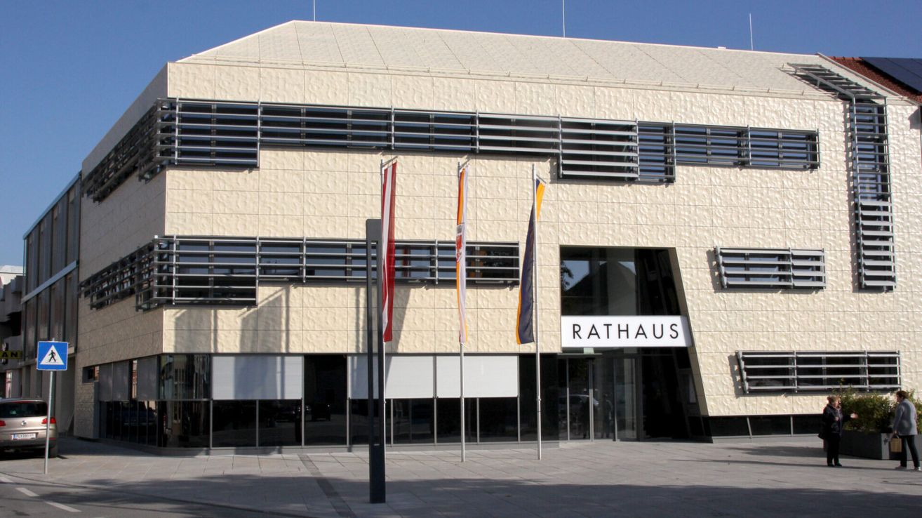 Die Vorderseite des Rathauses mit drei Fahnen davor. Das Gebäude hat eine geflieste Außenseite und Glasfenster. Das Wort Rathaus ist über dem Eingang angebracht.