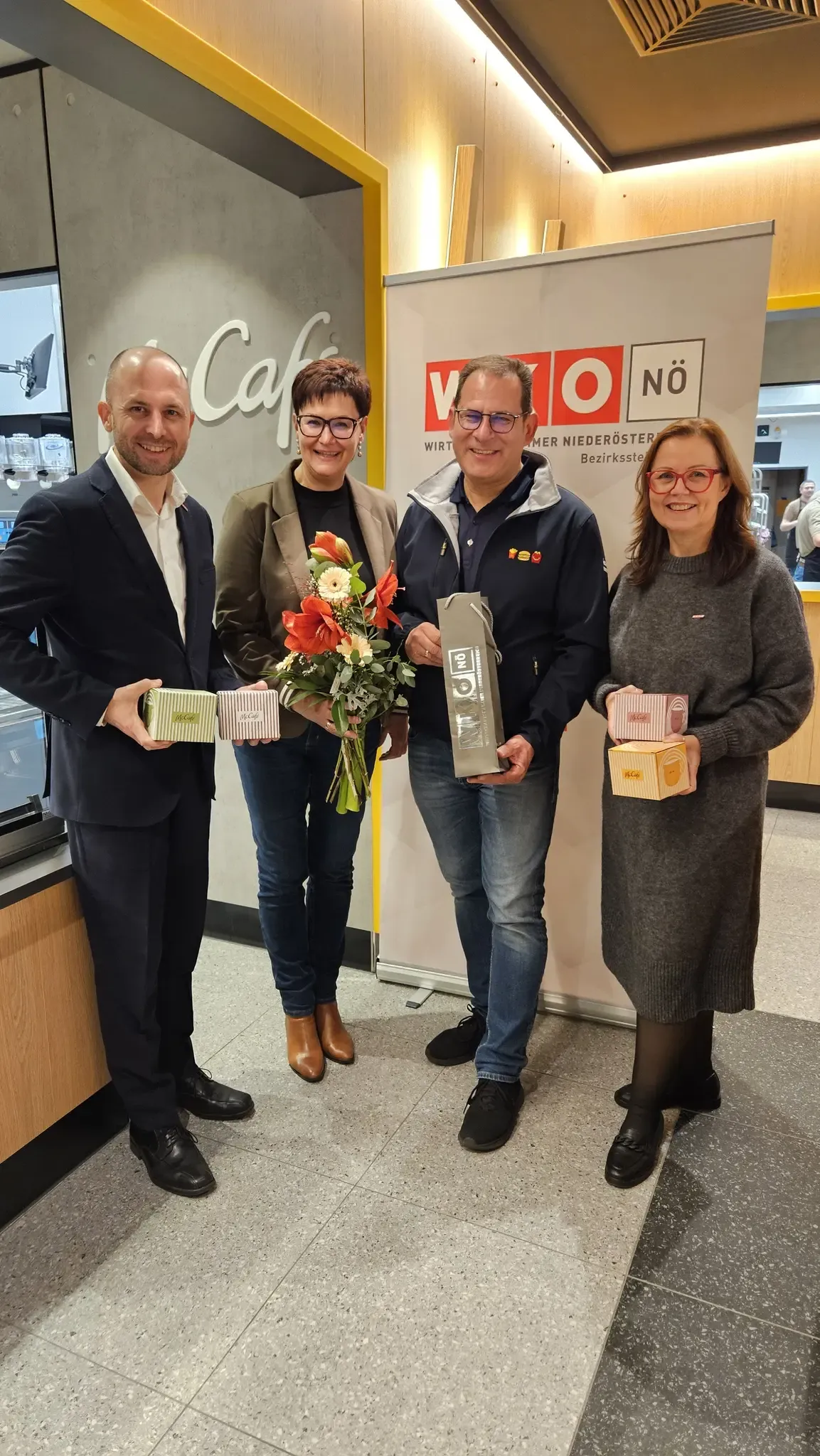 Vier Menschen stehen lächelnd zusammen in einem Café und halten Geschenkboxen. Der Mann links trägt einen Anzug, die Frau neben ihm hält einen Blumenstrauß. Dahinter steht ein Schild mit dem Wort 'Cafe' und einem Logo.