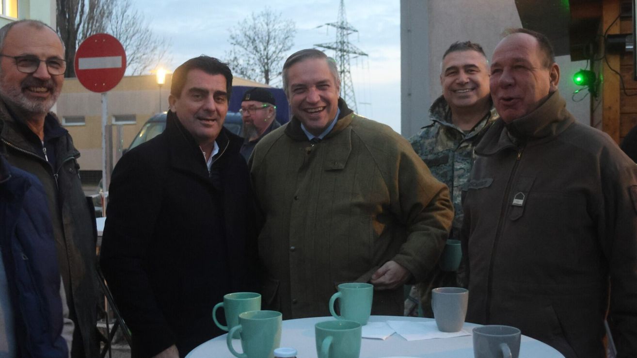 Fünf Männer stehen um einen Tisch mit Tassen, zwei lächeln. Einer in einer schwarzen Jacke, einer in einer braunen Jacke und drei weitere in Militäruniformen.