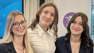 Drei Frauen posieren für ein Foto. Die Frau in der Mitte trägt ein weißes Hemd, während die anderen eine Brille und eine Halskette tragen. Hinter ihnen befindet sich ein weißer Banner mit einem lila Logo.