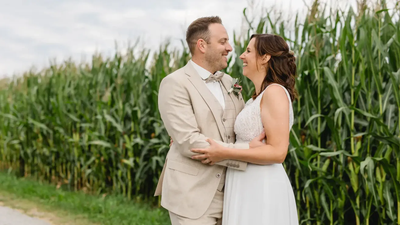 Ein lächelndes Paar steht vor einem Maisfeld, der Mann trägt einen beigen Anzug und eine Fliege, und die Frau ein weißes Kleid.