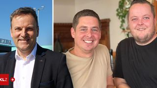 Drei Männer lächeln für ein Foto. Der linke Mann trägt einen schwarzen Anzug. Der mittlere Mann trägt ein beiges T-Shirt. Der rechte Mann trägt ein schwarzes T-Shirt. Der Hintergrund besteht aus einer hölzernen Wand und einer grünen Pflanze.