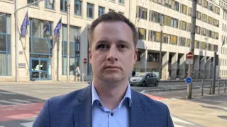 Ein Mann in einem blauen Anzug steht vor einem großen Bürogebäude mit einer Flagge und einem Auto auf der Straße.