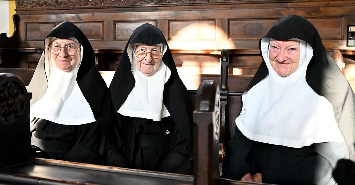 Drei ältere Nonnen in schwarzen Gewändern und weißen Schleiern sitzen auf hölzernen Bänken in einer Kirche. Sie lächeln alle und scheinen für ein Foto zu posieren.