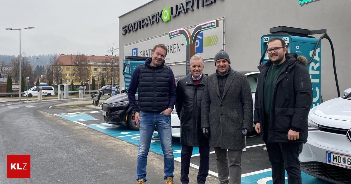 Vier Männer stehen vor einer Ladestation. Sie alle lächeln. Hinter ihnen befindet sich ein Gebäude mit dem Schild 'Stadpark Quartier'.