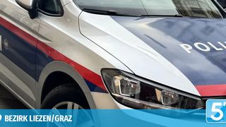 Vorderansicht eines Polizeiwagens aus dem Bezirk Liezen/Graz, mit roten und blauen Streifen und dem Wort 'POLIZEI'. Das Auto ist in einem städtischen Umfeld geparkt.