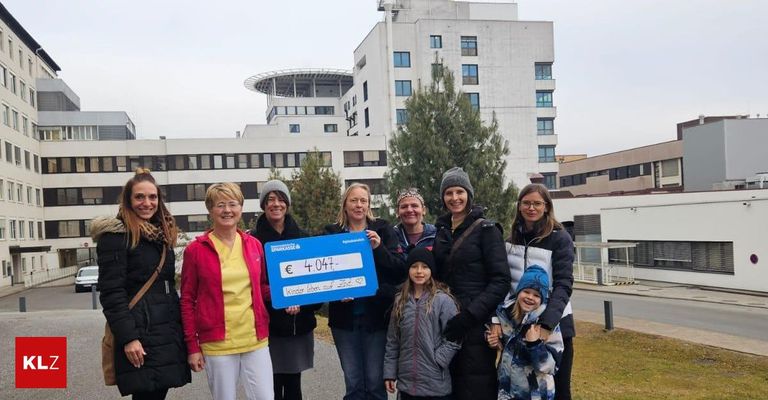 Eine Gruppe von Frauen und Kindern posiert für ein Foto vor einem Gebäude und hält ein blaues Schild mit der Aufschrift 'Spendenabrechnung € 4.047.' Dahinter steht ein hohes Gebäude mit vielen Fenstern und einem Baum.