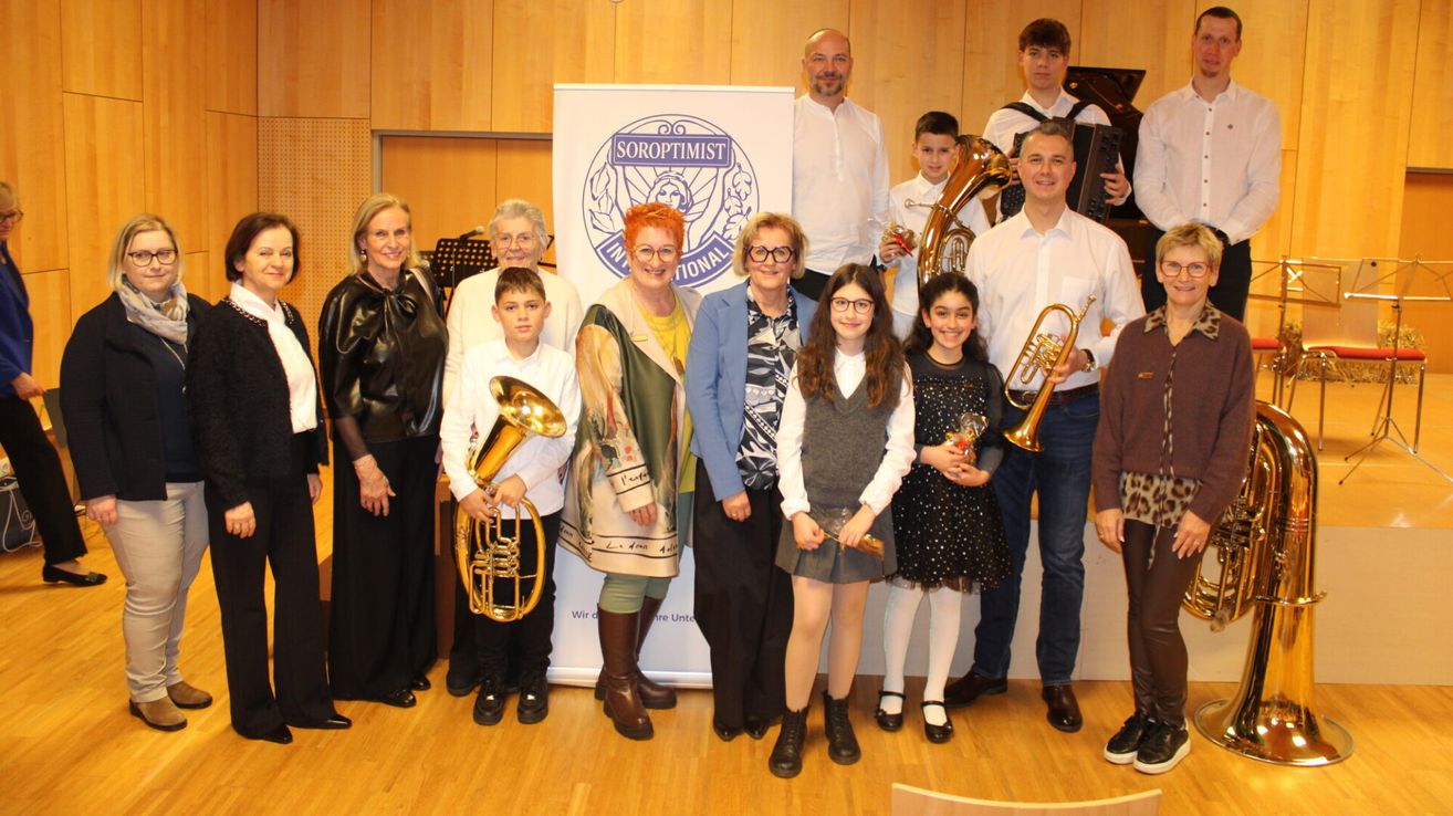 Eine Gruppe von Erwachsenen und Kindern posiert mit Musikinstrumenten, darunter eine Tuba und Trompete, vor einem Soroptimist International Banner.