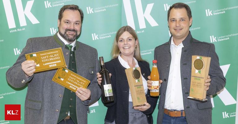 Drei Personen stehen eng beieinander und halten Auszeichnungen, Flaschen und eine Trophäe. Der Mann auf der linken Seite trägt einen grauen Mantel und hält ein Zertifikat. Die Frau in der Mitte hält eine Trophäe und eine Flasche. Der Mann auf der rechten Seite trägt einen grauen Blazer und hält eine Flasche Saft. Hinter ihnen ist ein grüner Hintergrund mit dem Text 'IK Landwirtschaftskammer Steiermark'.