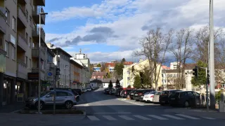 Eine Straße mit geparkten Autos auf beiden Seiten unter einem teilweise bewölkten Himmel. Bäume und Gebäude sind im Hintergrund sichtbar.