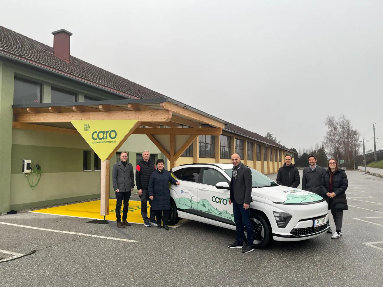 Eine Gruppe von Personen steht vor einem weißen Auto mit dem Wort Caro an der Seite, das auf einem Betonuntergrund geparkt ist. Sie tragen Winterkleidung. Hinter ihnen befindet sich ein Gebäude mit einem gelben Schild und einem hölzernen Vordach.